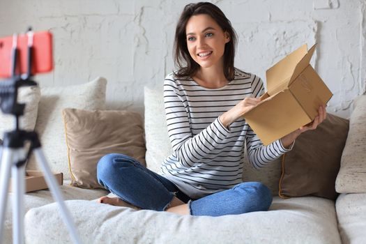 Young beautiful female blogger recording video while unpacking parcel at home