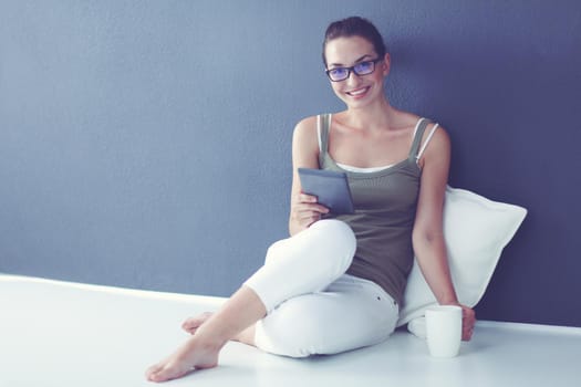 Beautiful young woman with cup of tea using tablet computer while sitting on floor at home.