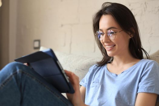 Smiling young woman sitting on sofa with digital tablet and chating with friends