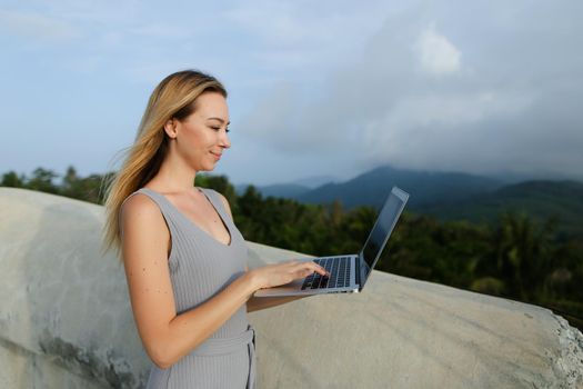 Young woman using laptop in green mountains background, wearing grey shirt. Concept of modern technology, summet vacations and nature.