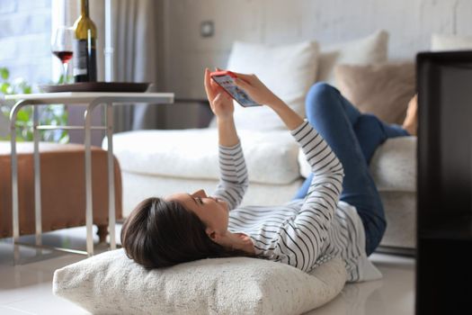 Young pretty woman checking social media, lying on floor against cozy domestic interior, drinking red wine
