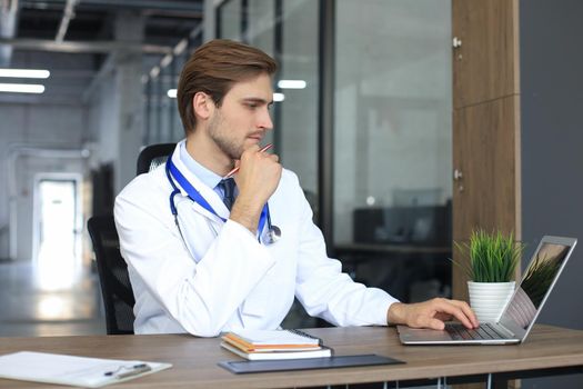 Doctor in white medical uniform with statoscope reading personal file of patient on tablet and writes data to laptop