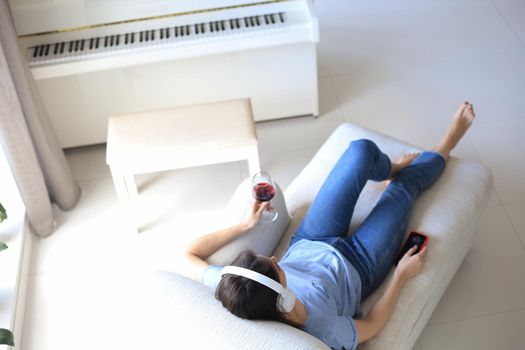 Beautiful brunette woman in earphones sitting on sofa listening to music with closed eyes, drinking red wine