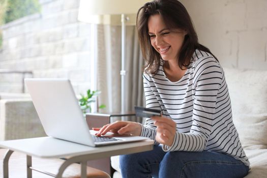 Happy natural brunette using credit card and laptop in the living room