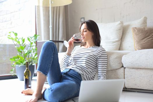 Smiling young woman sitting on floor with laptop computer and chating with friends, drinking wine