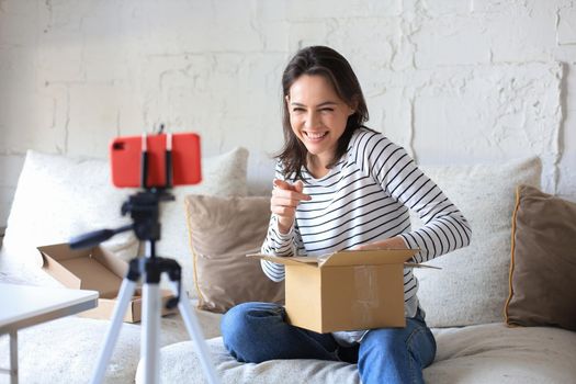 Young beautiful female blogger recording video while unpacking parcel at home
