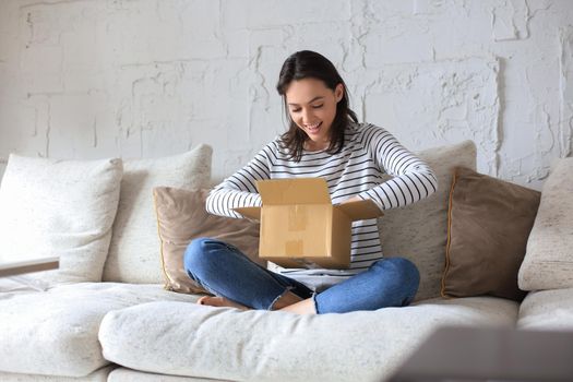 Beautiful young woman is holding cardboard box and unpacking it sitting on sofa at home