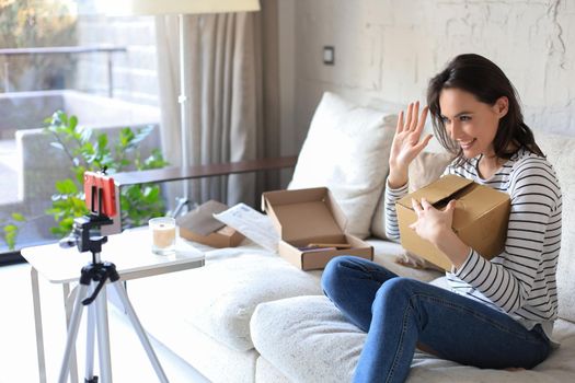 Young beautiful female blogger recording video while unpacking parcel at home