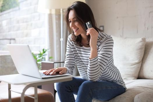 Happy natural brunette using credit card and laptop in the living room