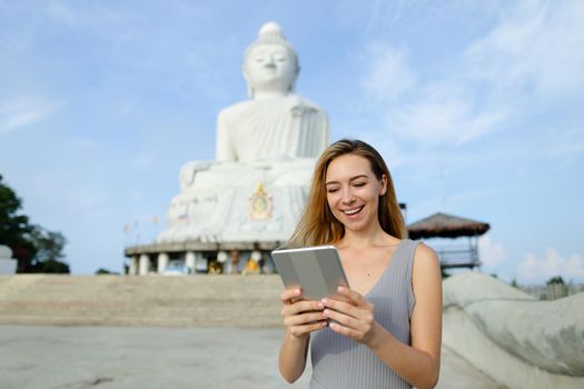 Young caucasian woman using tablet near white statue of Buddha in Phuket. Concept of traveling to Thailand and religion landmark, modern technology.