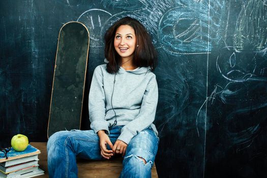 young cute teenage girl in classroom at blackboard seating on table smiling, modern hipster concept closeup