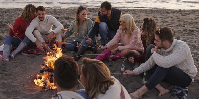 Group of young friends sitting by the fire late at night, grilling sausages and drinking beer, talking and having fun