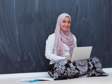 arab female student working on laptop from home  black chalkboard in background