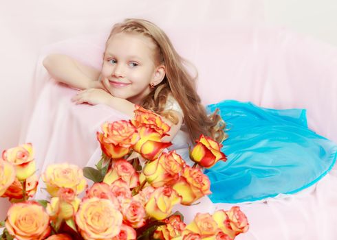 A beautiful little girl with long, light, curly hair, in a blue long skirt. She sits on a pink couch beside a large bouquet of tea roses.