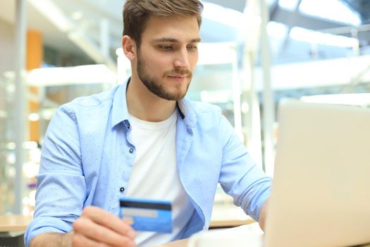 Smiling man sitting in office and pays by credit card with his laptop