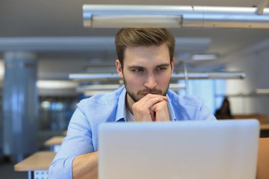 Young man using a laptop sitting thinking at his desk as he reads information on the screen