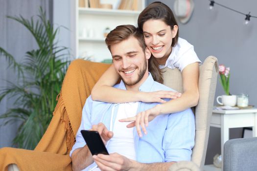 Young couple watching online content in a smart phone sitting on chair at home in the living room