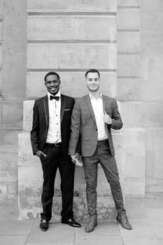 Black and white photo afro american and caucasian happy gays standing near building and wearing suits. Concept of lgbt and walking in city.