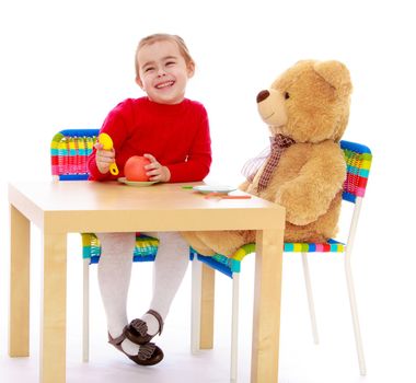 Cute little girl in a red dress in a Montessori kindergarten. Girl sitting at the table with a large Teddy bear and feeds him an Apple - Isolated on white background