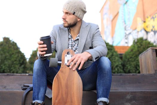 Handsome young man in grey coat and hat, resting, sitting with longboard drinking coffee. Urban skateboarding concept