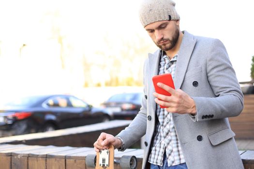 Handsome young man in grey coat and hat using smartphone, resting, standing with longboard. Urban skateboarding concept