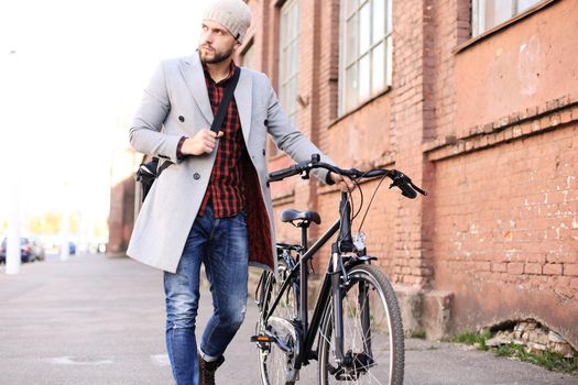 Handsome young man in grey coat and hat pushing his bike down a street in the city