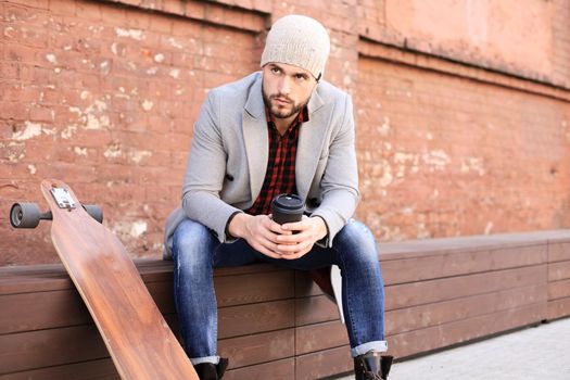 Handsome young man in grey coat and hat, resting, sitting with longboard. Urban skateboarding concept