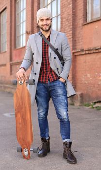 Handsome young man in grey coat and hat walking on the street, using longboard