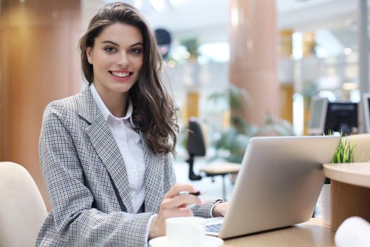 Portrait of a cheerful young businesswoman sitting at the table in office and looking at camera