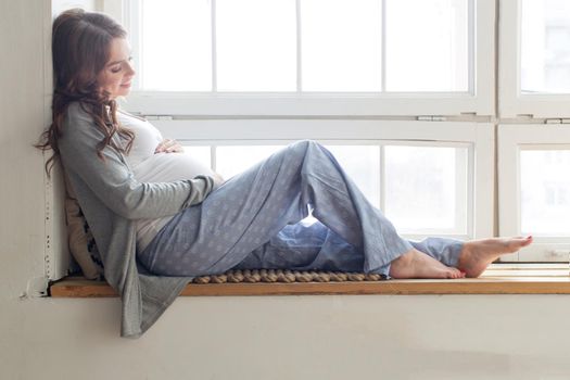 Pregnant smiling brunette female sitting on the windowsill touching her belly.
