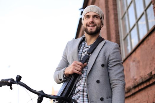 Handsome young man in grey coat and hat standing with his bicycle