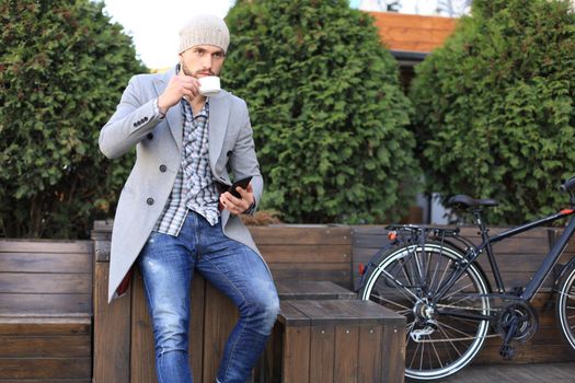 Handsome young man in grey coat and hat using smartphone, resting, and smiling while sitting near his bicycle outdoors