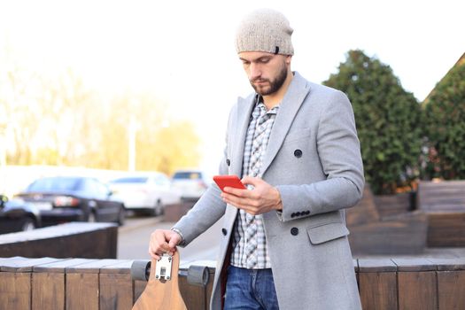 Handsome young man in grey coat and hat using smartphone, resting, standing with longboard. Urban skateboarding concept