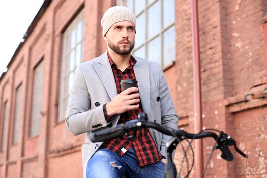 Handsome young man in grey coat and hat and riding a bicycle the street drinking coffee