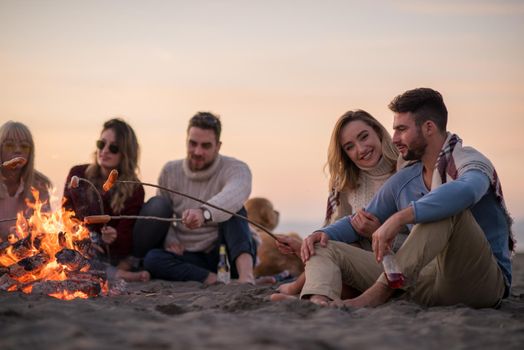 Group of young friends sitting by the fire at autumn beach, grilling sausages and drinking beer, talking and having fun