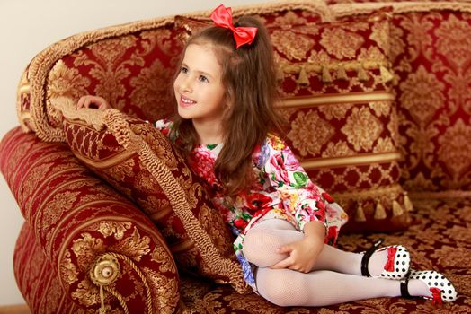 Nice little girl with a long ponytail down to his shoulders, and a red bow on her head, in a beautiful summer dress sitting on the couch. The girl looks away from the camera