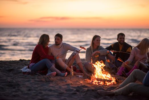 Group of young friends sitting by the fire at autumn beach, grilling sausages and drinking beer, talking and having fun