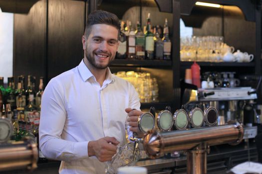 Handsome bartender is smiling and filling a glass with beer while standing at bar counter in pub
