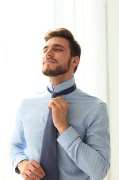 Morning dress up. Handsome young man in blue shirt adjusting his necktie and looking away