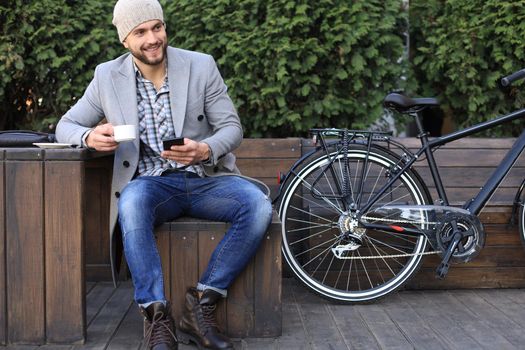 Handsome young man in grey coat and hat using smartphone, resting, and smiling while sitting near his bicycle outdoors
