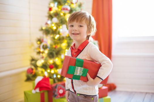 Little boy hold a gift box near a decorated Christmas tree at home.