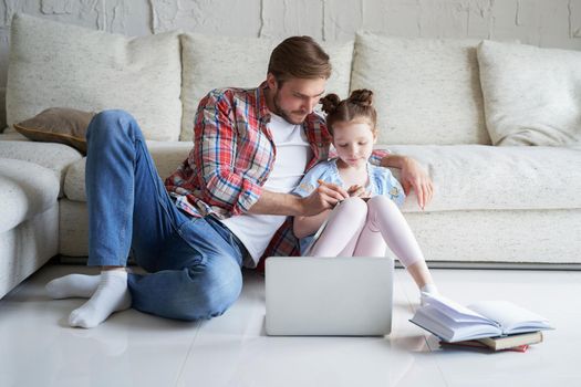 Smiling father and daughter sitting on floor in living room with laptop, teaching lessons