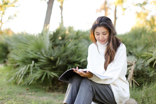 Japanese pretty female student sitting on stump in park and reading papers. Concept of preparing before exam and asian woman.