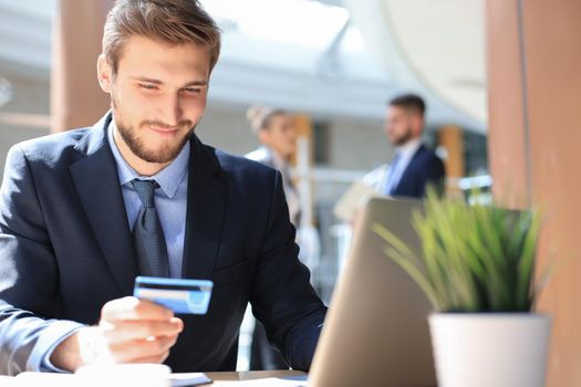 Smiling man sitting in office and pays by credit card with his laptop