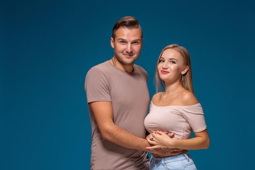 Young couple is hugging on blue background in studio. They wear T-shirts, jeans and smile. Friendship, love and relationships concept