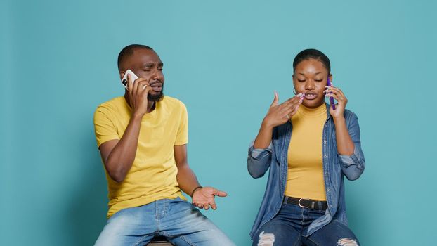 People having conversation on phone call while they sit together in studio. Man and woman using internet connection to talk on mobile phone communication. Modern couple with smartphone