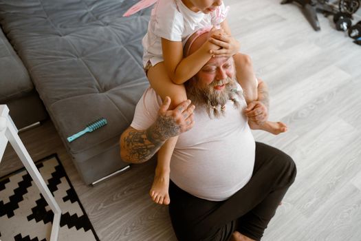 Little daughter sits on shoulders of positive plump father with scrunchies in beard near grey sofa on floor in living room. Family playtime