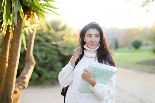 Japanese girl with documents walking in park and wearing white sweater with backpack. Concept of asian student.