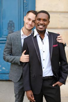 Two smiling boys, caucasian and afro american, wearing suits standing near building and hugging. Concept of gays and lgbt.