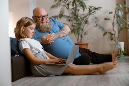 Smiling dad with glasses and cute little girl watch video via contemporary laptop sitting on floor in bedroom. Device for fun and work